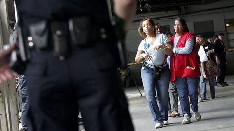 A line of people in pairs, at the front of the line, a person in a red apron leads a person who appears to be injured.