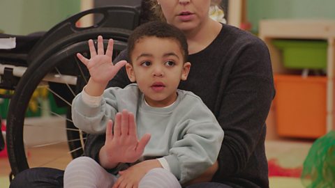 A son and his mum sit in the toddler club and hold up their hands to indicate "stop".