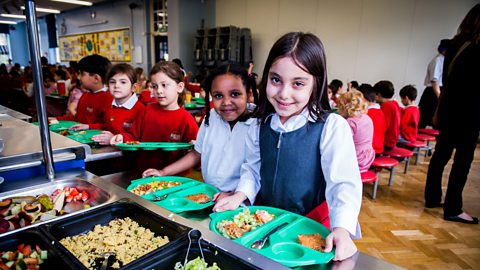 A dinner hall in a UK primary school. Pupils are holding their lunch trays and smiling.