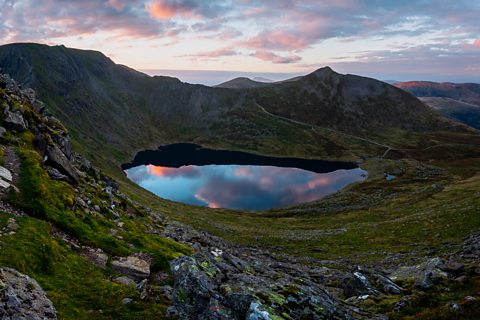 An image of a small lake on the side of a hill with some steep edged and some lower edges