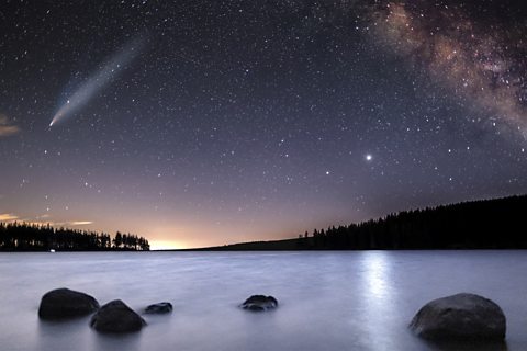 An image of a comet with a long 'tail' travelling over the night sky above a lake.