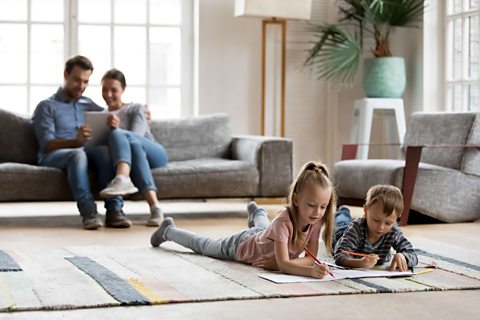 Two adults look at tablet on the couch as two children draw on the carpet.