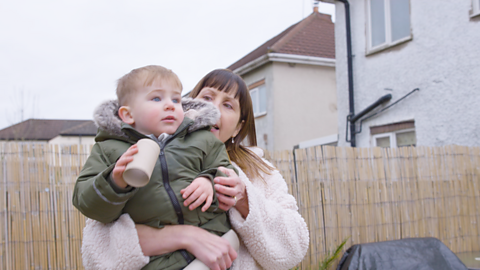 A grandparent and her grandson are out in the garden playing with a cardboard telescope. She is carrying him and he is looking at the sky.