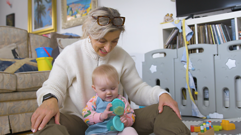 A grandma and her young granddaughter since in a living room and play with a homemade shaker or rattle.