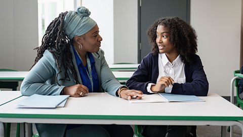 A student and a teacher sitting behind a white desk. The teacher is showing the student something from a book.