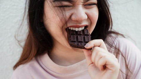 Picture of a young woman eating a bar of dark chocolate and looking happy.