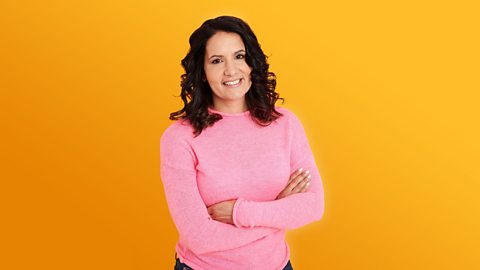 Dr Radha standing in front of a yellow background. She is wearing a pink sweater and is smiling with her arms crossed.