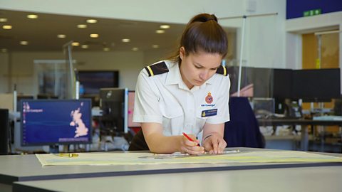 A picture of a young lady named Sydney, with brown hair, wearing a white shirt and plotting on a paper map with a pencil.