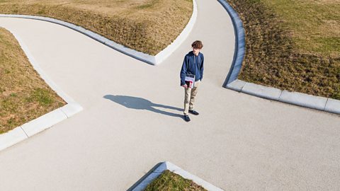 An image of a boy holding a textbook, standing in the middle of a path that splits into four directions