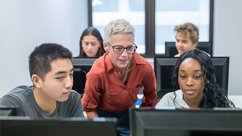 An image of young people on computers, with an older lady looking over their shoulder at the screen