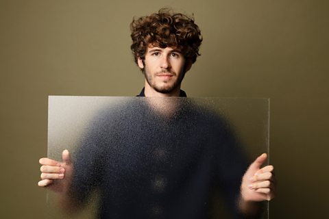 A person holding a sheet of frosted glass in front of them - Their head and hands that are no behind the glass appear clear, the body of the person appears blurred behind the frosted glass.