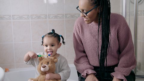 A little girl brushing her teeth as her mum looks on.