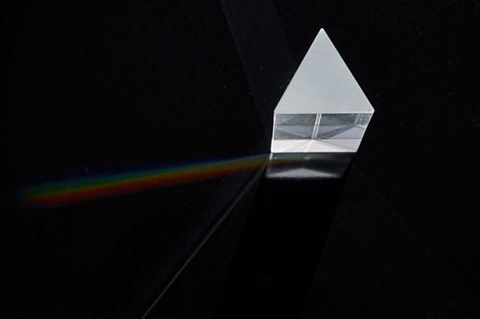 A triangular prism on a black background . A single ray of white light comes out split into the spectrum of the colours of the rainbow.