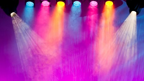 A photo of different colours of stage lights beaming down onto a smoky stage.