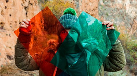 A woman stood in the wilderness holding up a large piece of red and a large piece of green cellophane in front of her - It makes the images behind her appear different