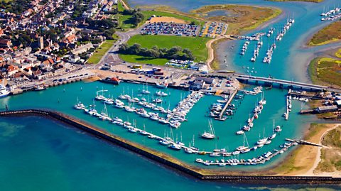 A an aerial photo of the harbour in Yarmouth, with a stone wall, aqua water and moored white sailing boats.