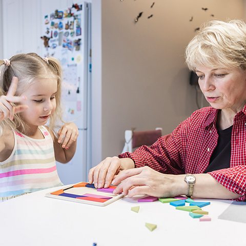 Focused little girl plays with tangram puzzle at dinner table in the kitchen with her grandmother. 