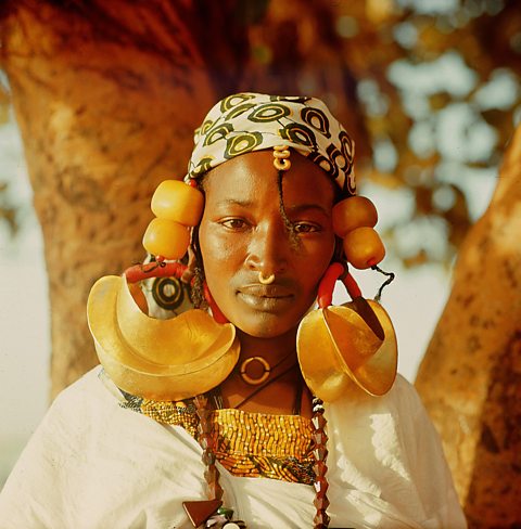 A black woman wearing a white and green head covering and a white dress. She is wearing large gold crescent earrings and large amber beads in her braided hair.