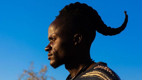 A photo of the side profile of a black man against a blue sky, wearing a woven shirt. He is hair is long and style in a single plait, with a curve to it.