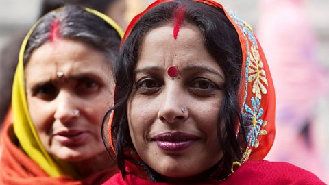 A photo of two Indian women wearing traditional sarees, one orange and one yellow. Both have red sindoor powder applied to their parting and one is wearing a red bindi.