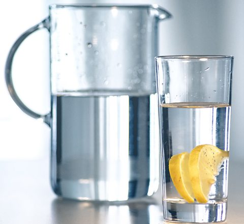 An image of a jug with clear water allowing light to pass through clear liquid in a clear cup that contains lemon slices into a glass