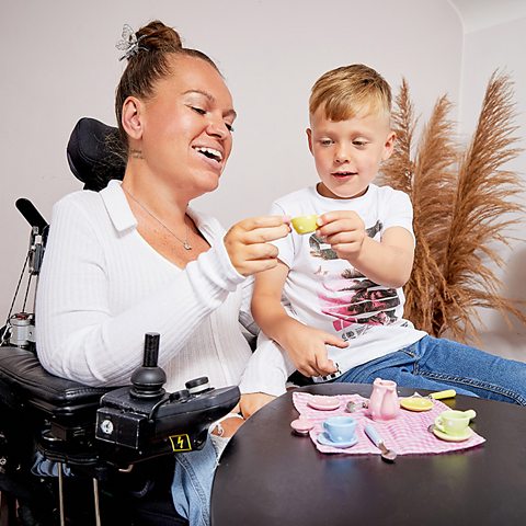 A mum and her son play with a tea party set. The mum sits in an electric wheelchair and her son sits on her lap. The tea party set is on the table.