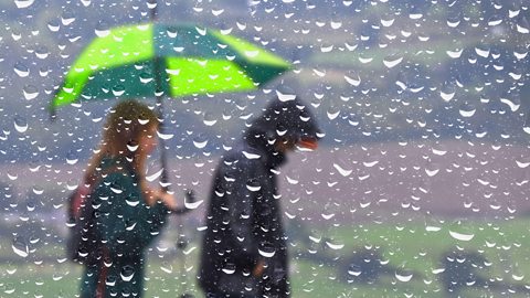 A couple walk in rain with an umbrella, seen through a window covered in rain droplets.