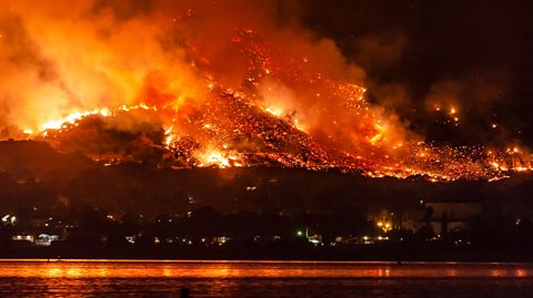 A photo taken at night of a wooded hill on fire, with a lake in the foreground reflecting the orange light.