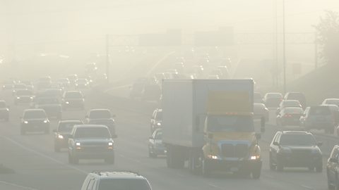 A photo of heavy traffic on a motorway with thick smog. Many of the vehicles have their headlights switched on.