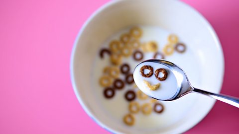 A bowl of cereal over pink background, with a spoon floating over it containing milk and a happy face made of cereal.