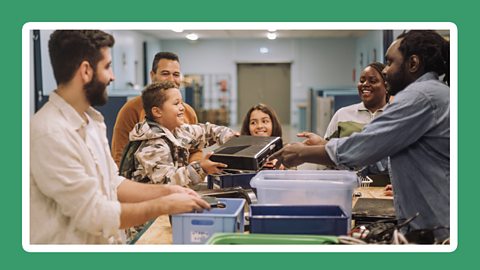 A child handing over an electronic device to a man in a community centre while others watch