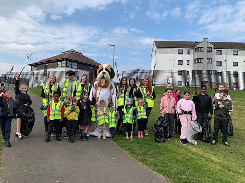 Group of children stand with their school mascot, a big dog, and litter pickers. 