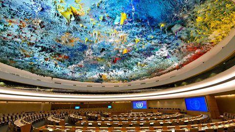 A huge circular meeting room. The ceiling is covered in thick layers of paint, mainly blue, yellow and red. In places, the paint hangs down in large drops, giving a textured effect.