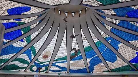 An enormous ceiling of stained glass, supported by curved concrete pillars which meet a point at the centre of the ceiling. Wavy bands of blue, green and brown glass stretch out across the ceiling.