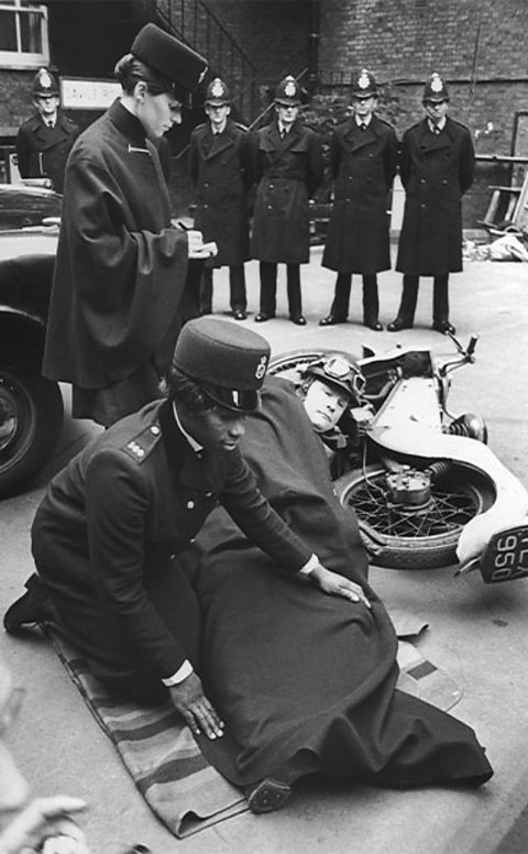 A female police officer covers an officer lying on the floor next to a motorbike with a blanket. A female officer takes notes and five male police officers stand at the back