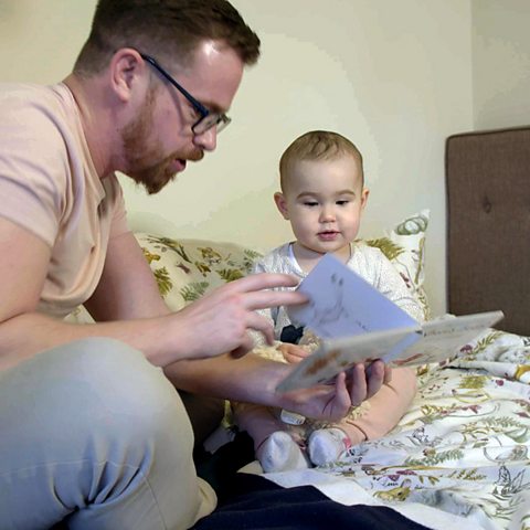 A dad and his daughter sit on a bed and look at a book together.