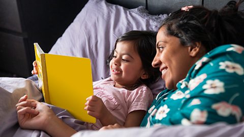 A mid adult woman and her daughter lying down in bed and reading a book together. It is the morning and they are both wearing pyjamas.