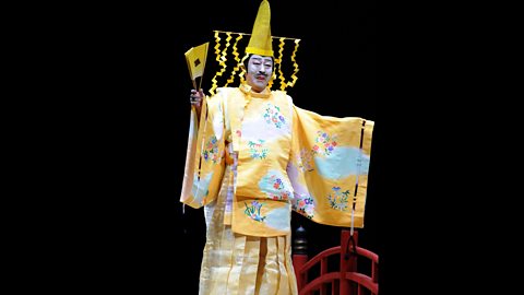 A Japanese actor wears a flamboyant yellow costume complete with a bright yellow fan and yellow headdress.