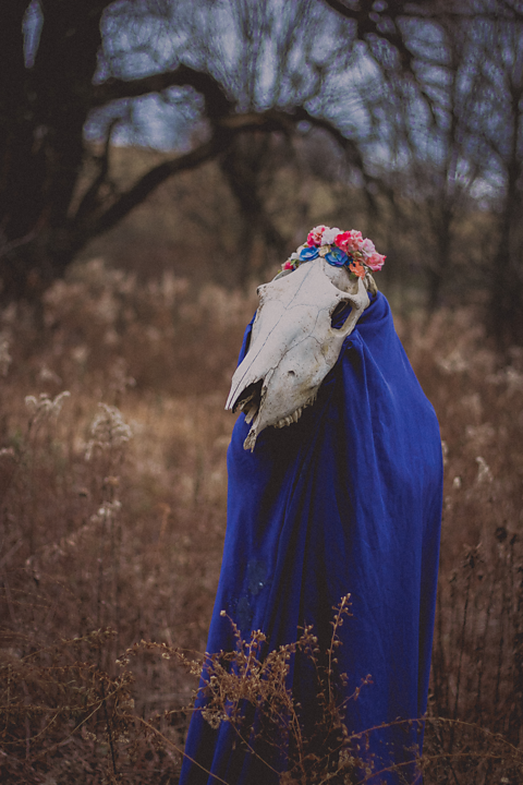 Someone dressed in a blue cloak and wearing a skeletal mask stands in the woods