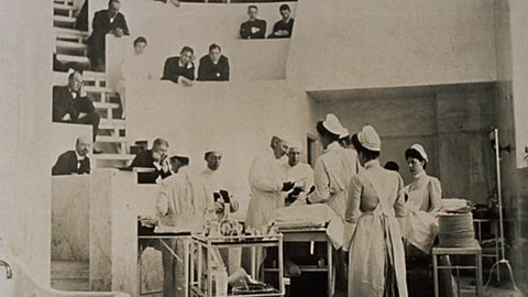 A black and white photograph of three doctors performing an operation with female assistants in the foreground and people watching from above.