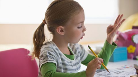 A young girl counts to four on her hand while writing with a pencil.