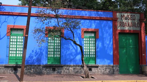 A photo of the front of Kahlo's house, which is a bright blue with a bright green door and windows and a small tree in front of it.