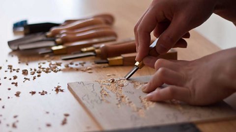 Closeup of a craftsman's hands carving a flat panel of wood with a specialist tool