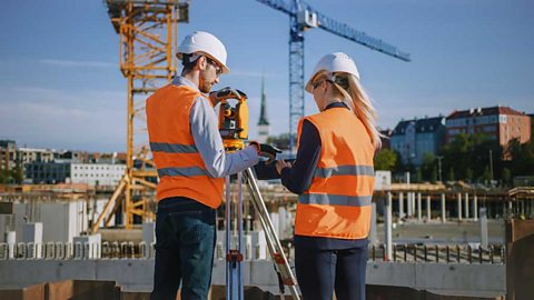 A man and woman in hardhats and orange hi vis jackets, a surveyor's tool on a tripod