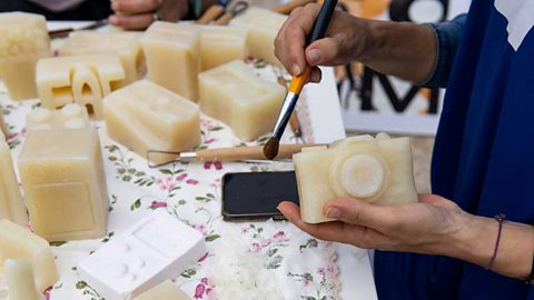 Hands holding a paintbrush and a soap carving of a camera. More  soap carvings on a table