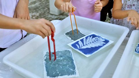 Children dipping plastic tongs into a tray of liquid and paper with artistic designs