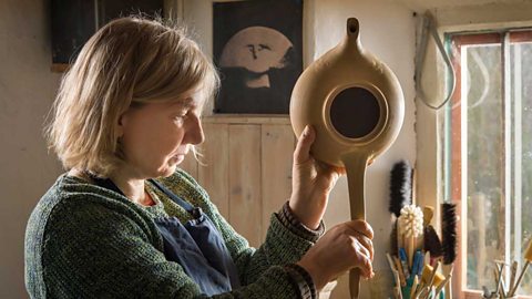 A woman in an apron working on a clay teapot by pulling a length of clay to form a handle