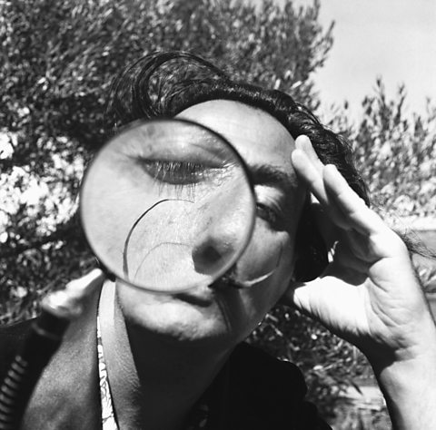 A black and white photo of Salvador Dali holding a magnifying glass in front of one eye