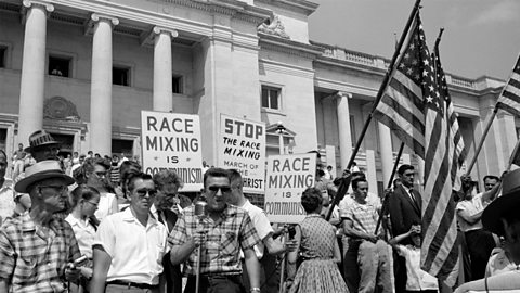 A group of protestors outside of a building holding signs that say things like 'stop the race mixing' and holding large American flags