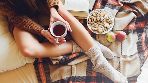 Person sits on a rug with hot drink and plate of cookies next to them, the person is wearing cosy socks